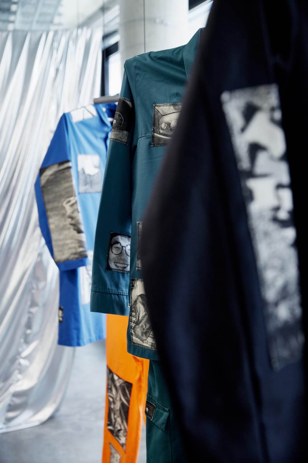 A close-up photograph of garments hanging in an exhibition. The black item in the foreground and blue jacket in the background are not in focus. In the centre of the image is a green jacket which has been adorned with black and white patches of photographs. The one in shot is a close up of a man in glasses, smiling. Behind the green jacket, the leg of an orange garment is visible.