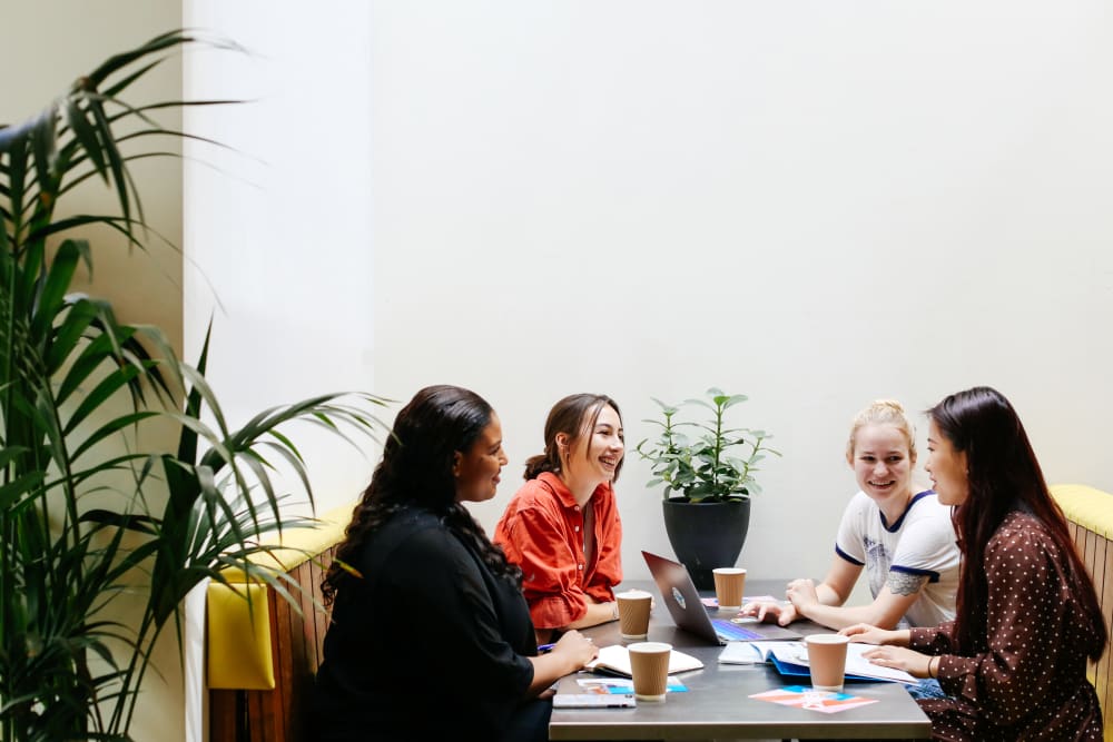 A group four women sit around a table with coffees and a laptop, talking and smiling. There are two plants in the room.