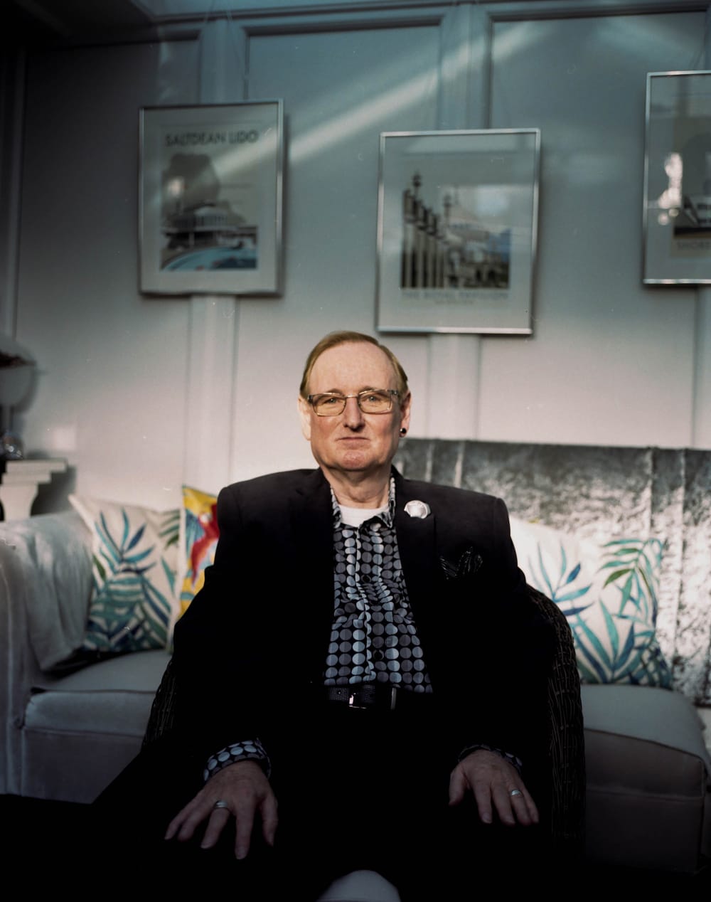 A white man in his 50's sitting facing the camera. His hands are rested on his lap he is wearing a suit with a checked shirt unbuttoned at the top. He is wearing glasses. Behind him a grey velvet sofa with cushions on. On the wall behind there are 3 frames each with photo but are slightly out of shot to describe. The wall is wooden with a grey paint.