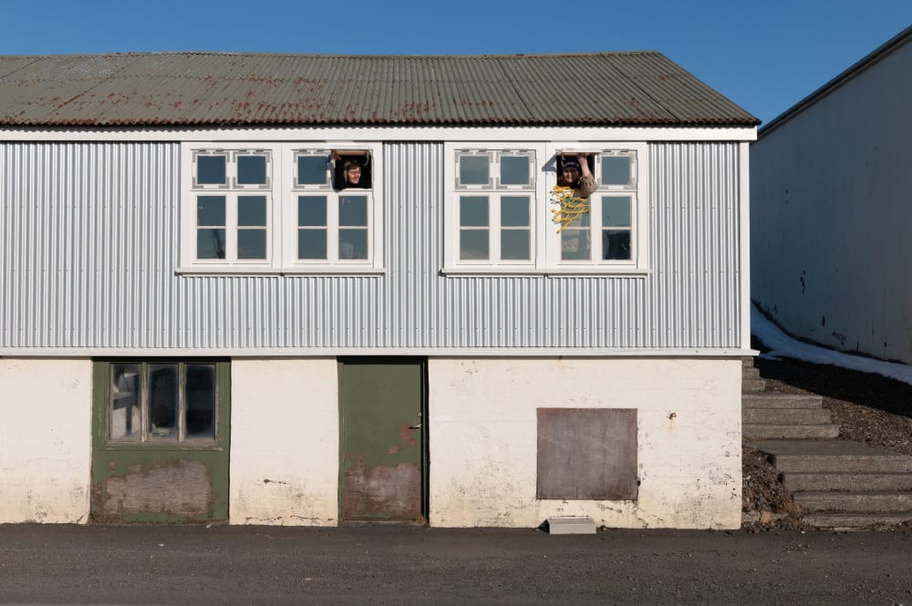 Two women looking out of small windows high up in the front of of a pale-coloured, functional-looking building.