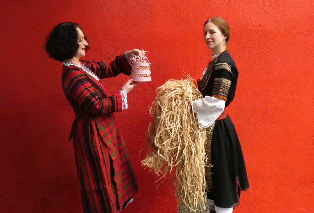 Two women in ethnic dress on a red background, one of them holds a pink bag