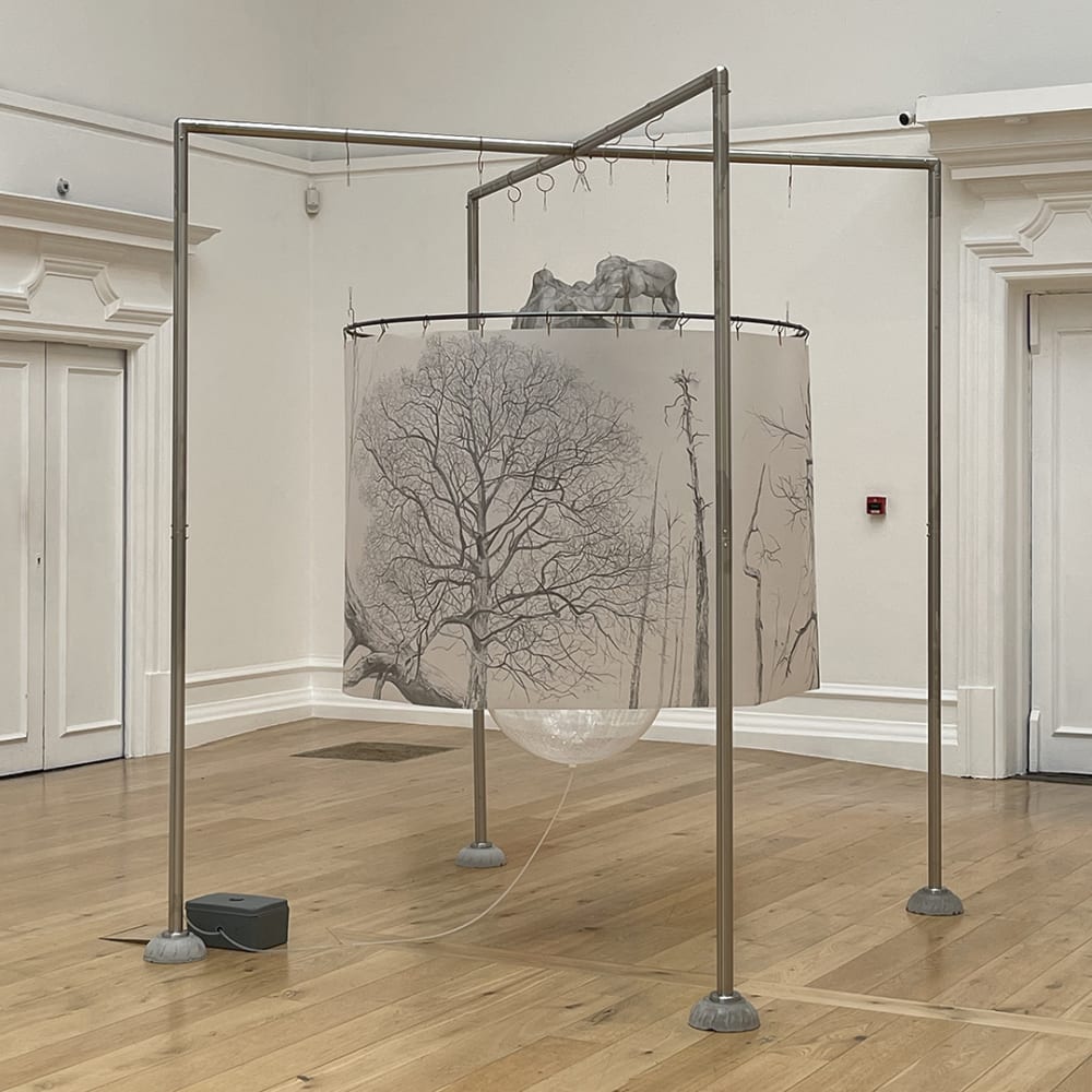A sculpture structure is shown in full. A metal frame is suspending the installation piece, showing a line drawing of trees hanging from a circular frame. 