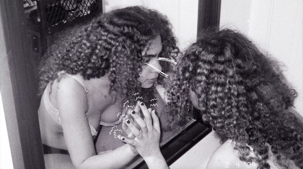 A black and white film photo of a person crouched in front of a mirror with their hands against it - they are staring at themselves. There are white doodles of zigzags around their fingers and two connecting lines between their eyes and their reflection's eyes.