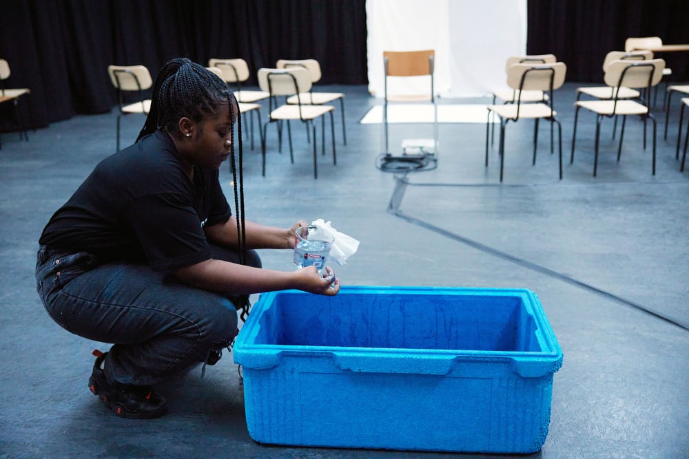 Image shows a young person in a studio with black flooring and light wooden chairs in the background. The person sits on the left hand side and is crouched over a blue box in which they are mixing blue paint