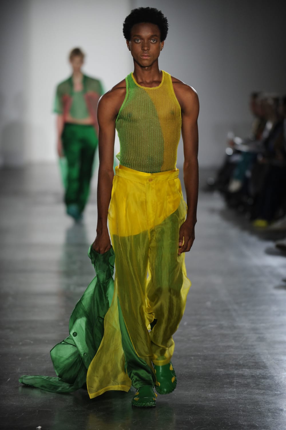 Man on runway wearing green and yellow netted vest paired with green and yellow silk pants with yellow chiffon slit overlay and matching green and yellow shoes, holding a matching green jacket dragging on floor with right hand 