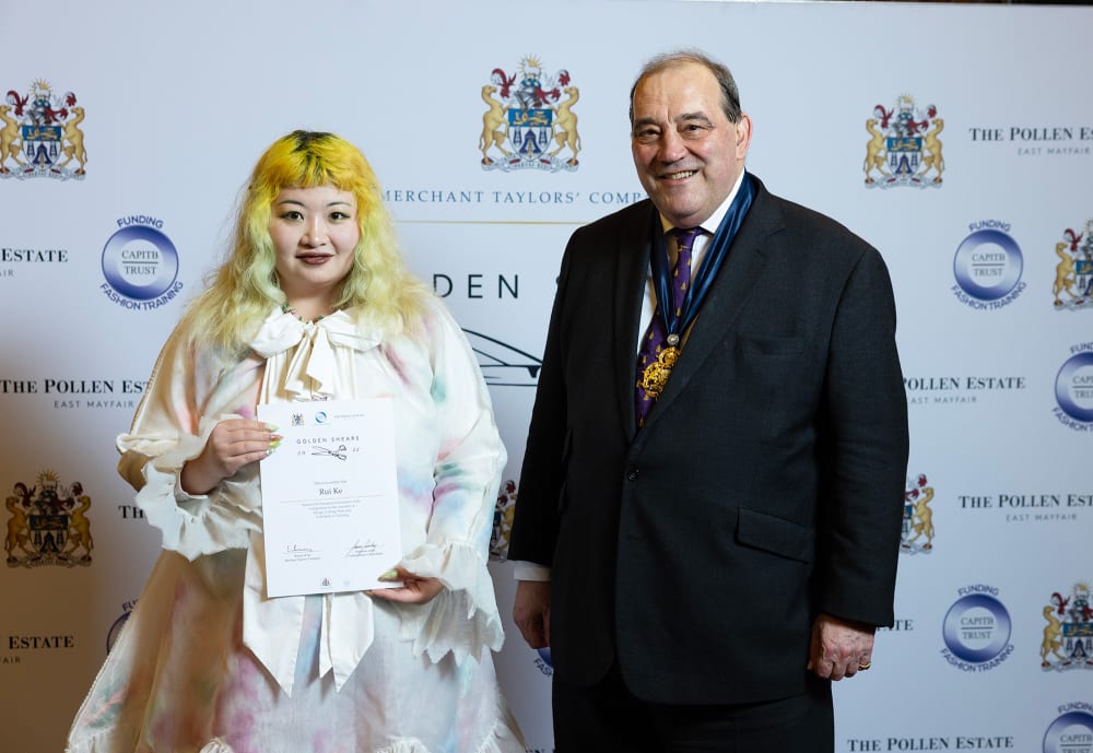 Image shows a person with yellow hair wearing a white top holding up a certificate and smiling at the camera. There is a person next to them in a black suit, also smiling at the camera.