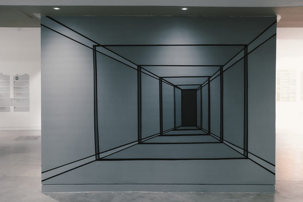 Image shows a wall with a long corridor painted in different shades of grey. The artist has used perspective to make it look as though you could walk into the corridor. There is a small black door at the end.