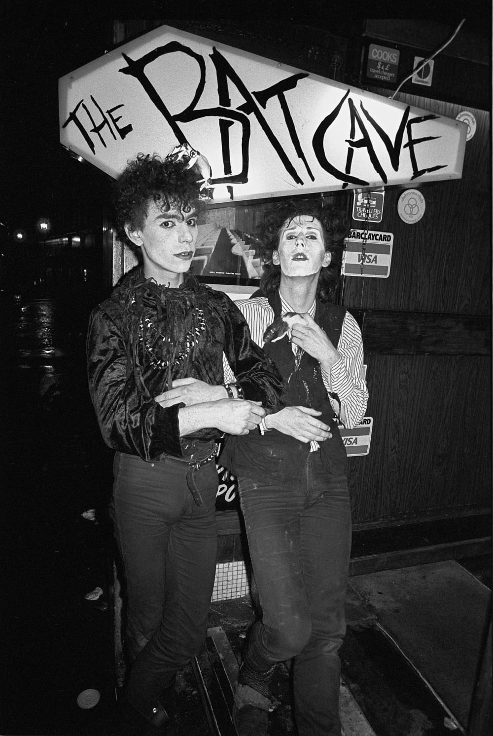 a black and white photograph of two goths under a sign that says BatCave