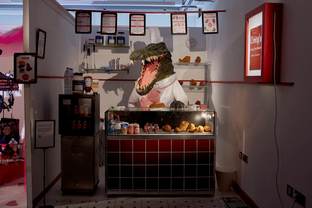 Image shows a model of a crocodile standing behind a model of a chicken shop. The crocodile has an apron on as if it works in the chicken shop and menus can be seen hanging from above the crocodile’s head.