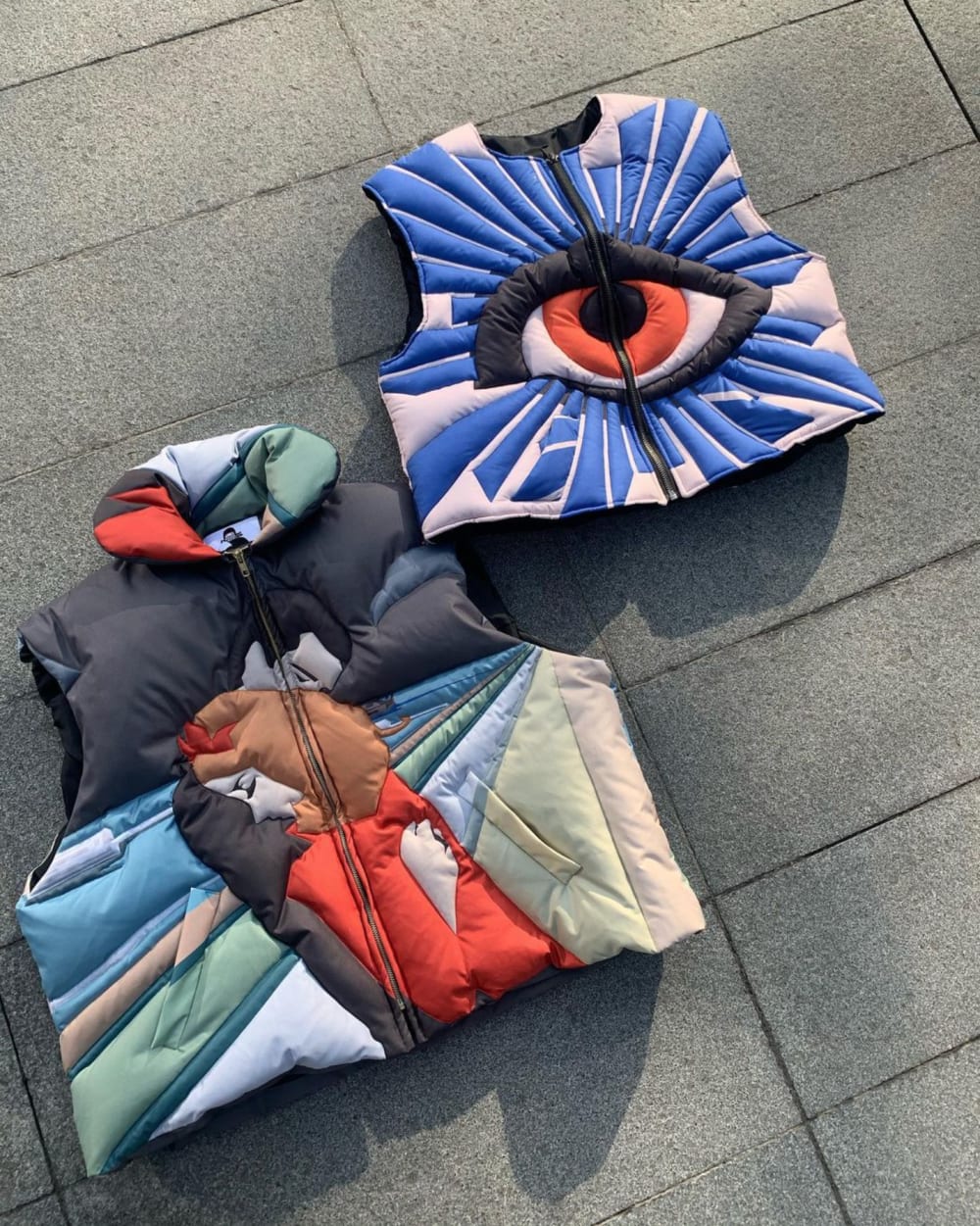 Two vests are placed on the ground; colourfully decorative one vest has an all-seeing eye on and the other has two people embracing. 
