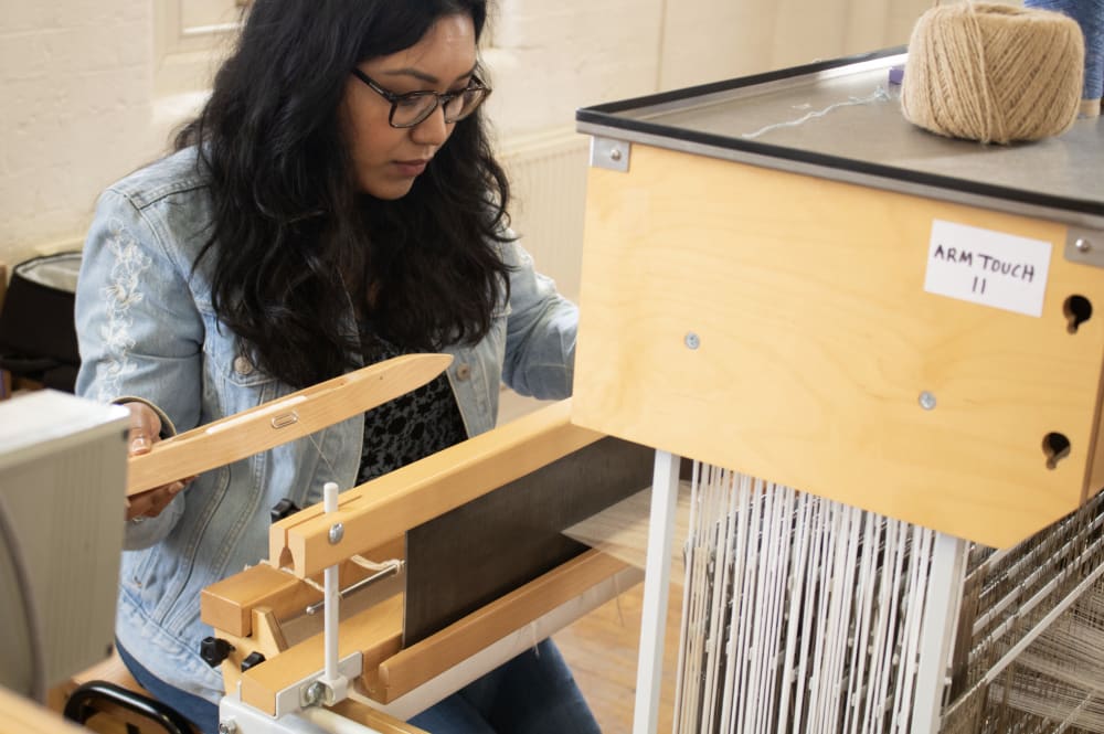 Anika with long dark hair, is sat at a loom weaving. 