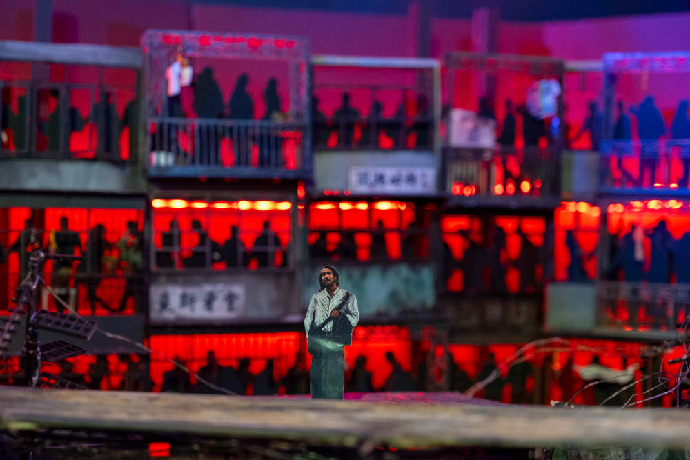 Image shows a small model figure standing in a set model box with red lighting and a crowd of people watching them from behind