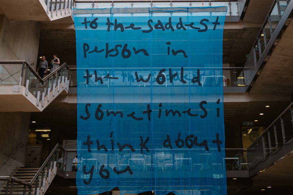 Large scale blue, semi-sheer artwork hanging from the second floor balcony. It reads: 'to the saddest person in the world sometimes I think about you'. 