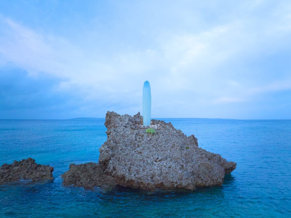 Image shows a large and thin blue object sitting on top of rocks in a blue ocean, with a light blue sky background.