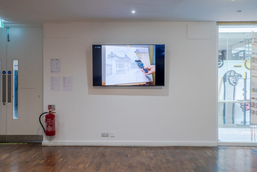 Screen hanging on a wall. On the screen is a hand drawing an intricate illustration of a building. 