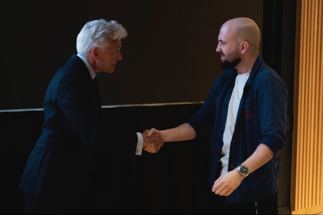 Two men shaking hands on with grey hair and a navy blue suit and the other with a bald head and navy blue shirt with a white t-shirt under it and black pants. 