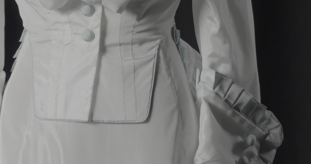 Close-up of a white, satin-like dress featuring structured seams, large buttons, and intricate ruffled details on the sleeves, conveying elegance and sophistication.