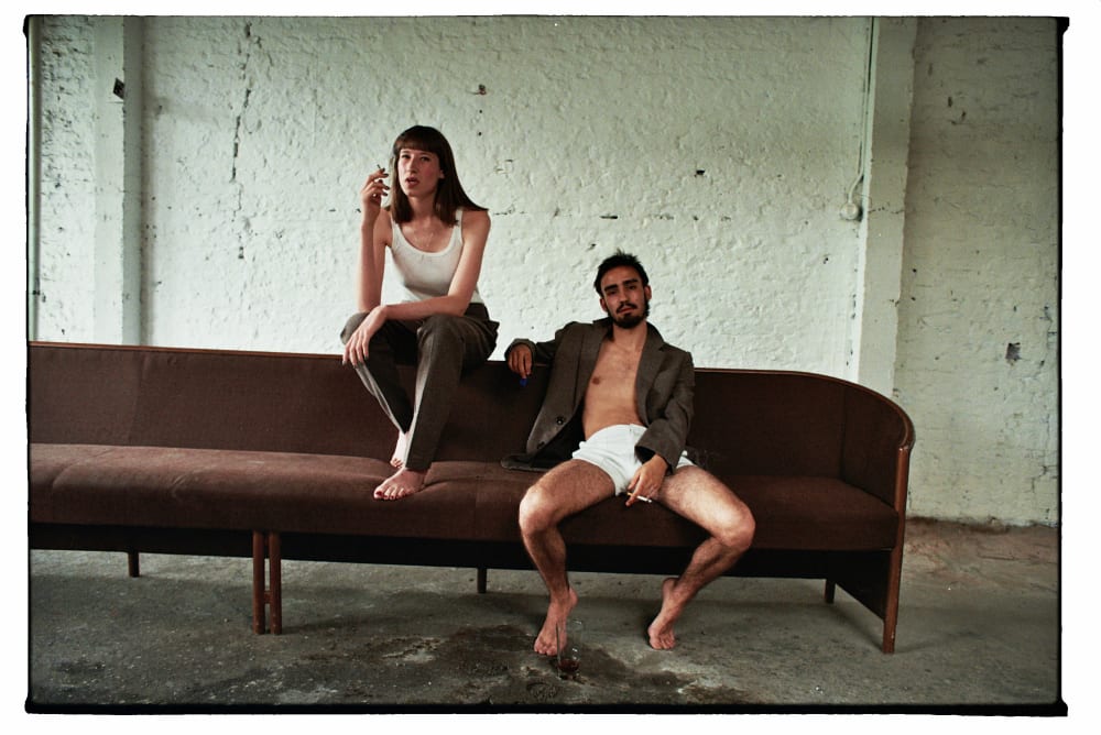 Two people sit on a brown couch in a minimalist room with a textured wall. One wears a tank top and pants, the other a jacket and shorts, both holding cigarettes. Moody vibe.