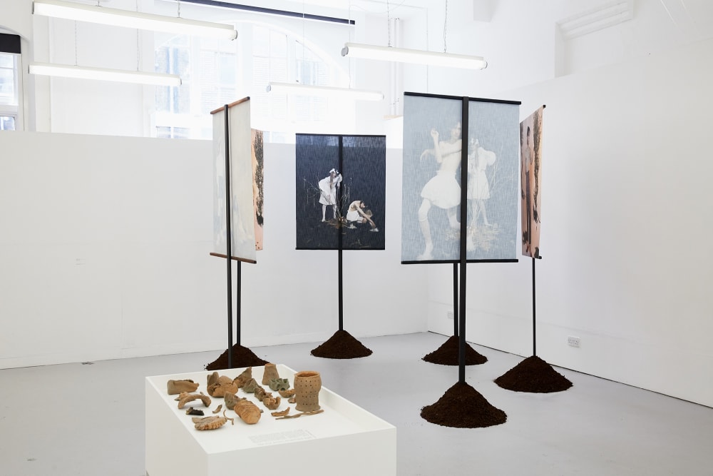 Installation shot of six banners on stands in a white exhibition space. The banners show photographic images of people. In the foreground is a low plinth with small objects on it.