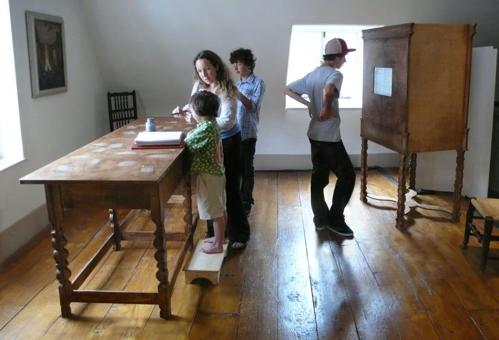 A woman and two children work at a high table in a room with wooden floorboards, while a young man in a hat reads a noticed pinned to a wooden cabinet behind them.