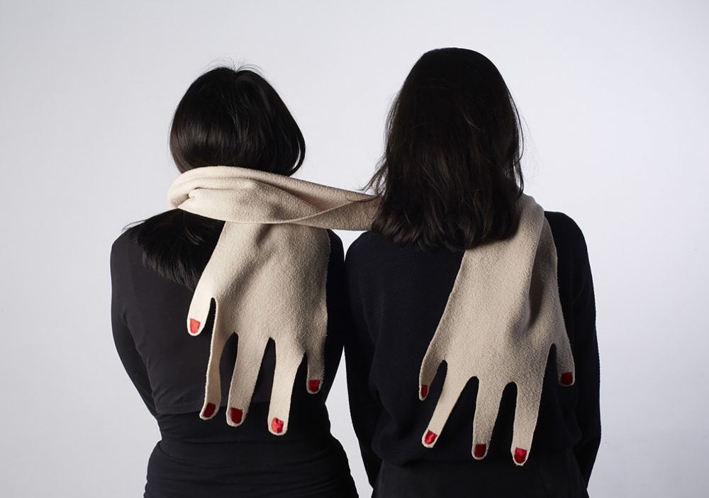 Two Camberwell BA Graphic Design students sharing a giant scarf that is designed to look like a pair of giant hands.