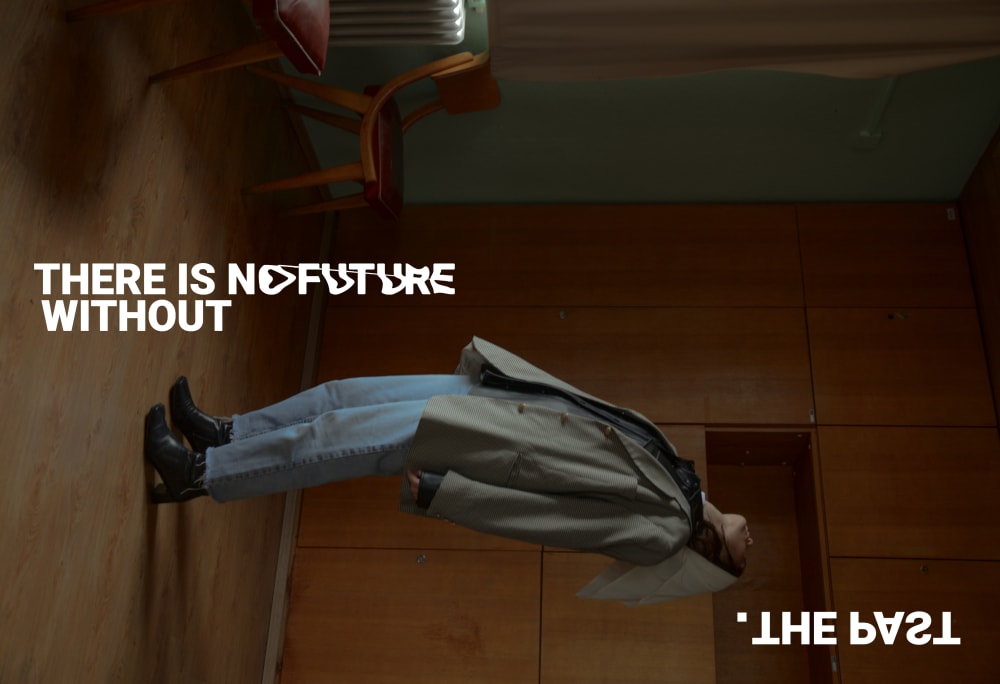 sideways photo, model leaning backwards, wearing overcoat, jeans, millinery and boots. Stylised caption reads