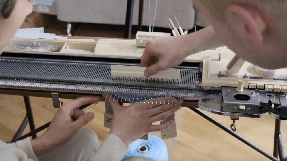 A close-up image of two people using a knitting machine. One person is holding the knitted piece in place while the other person is holding a part of the knitting machine above.  