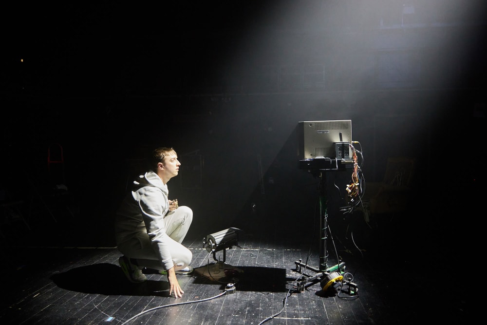 A man dressed in white crouches down on a stage, looking up at a tv monitor on a tripod.