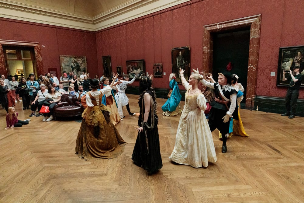 A group of performers in different costumes stand in a circle in a large grand gallery space in front of a crowd of people.