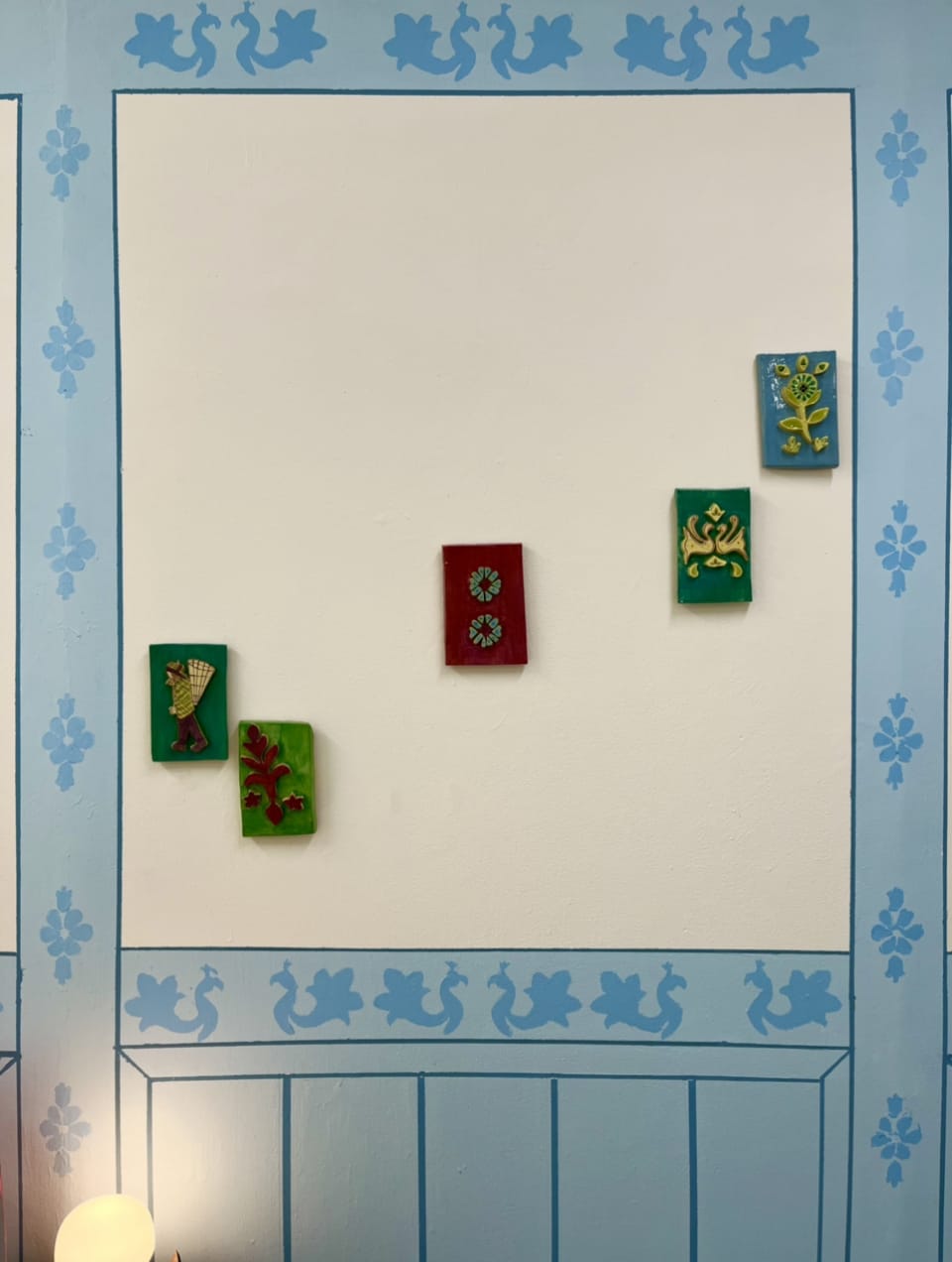 A wall with a blue decorative boarder and a square cream centre where 5 colourful rectangle shaped ceramics with individual designs are hung sporadically. 