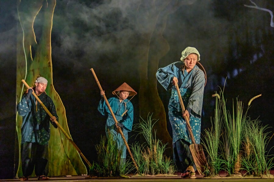 Image shows 3 people wearing traditional Japanese clothing holding agriculture tools amongst reeds 
