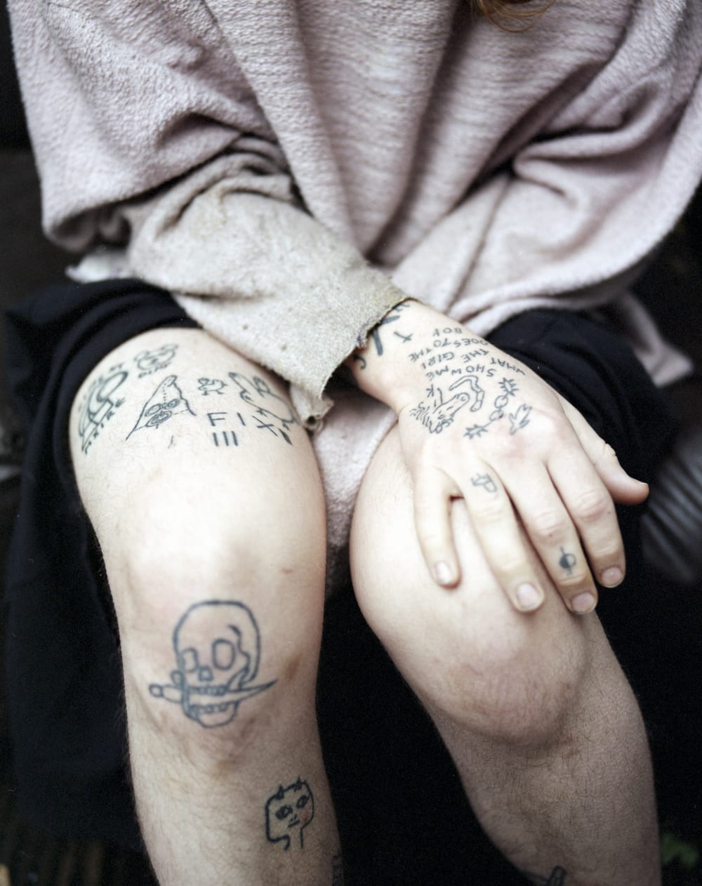 A hand rests on a knee. Both hand and knees are tattooed.