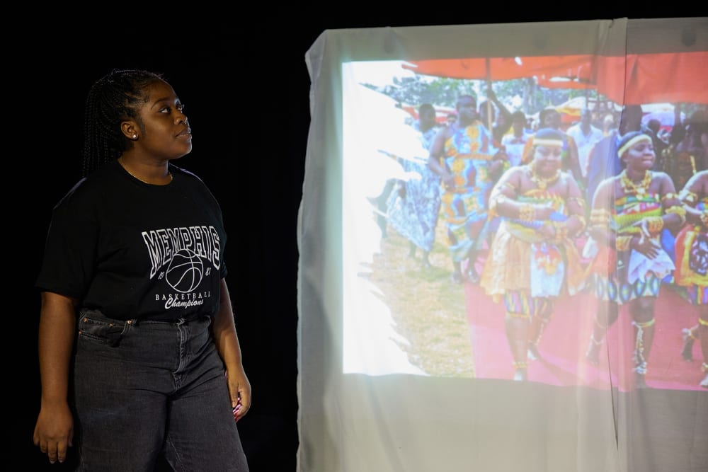 Image shows a young person on the left hand side looking at a projection of traditional dancers from Ghana. The dancers are wearing traditional dress.