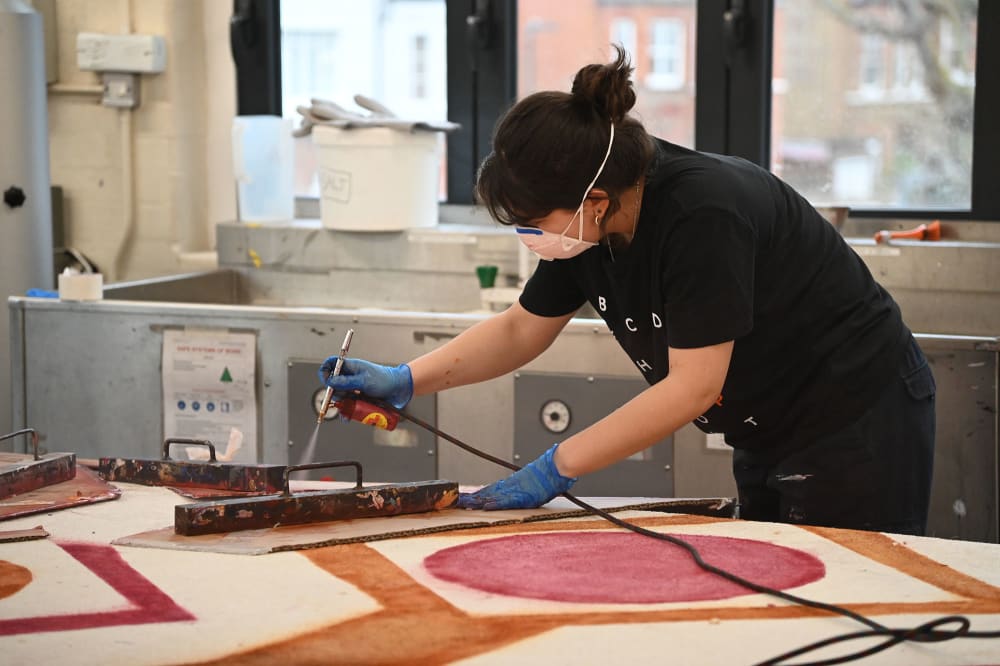 Image shows a person in a painting lab with a mask on spray painting carpet to create a 1970s inspired rug. The person is wearing all black with their hair tied up and light windows sit behind them.