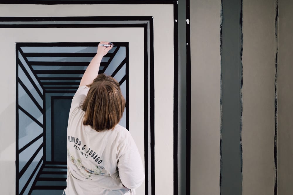 Image shows a person in a white top and short brown hair holding a pen and reaching up with their left hand to draw a line on a wall. The wall itself contains a half-completed artwork using different shades of grey paint