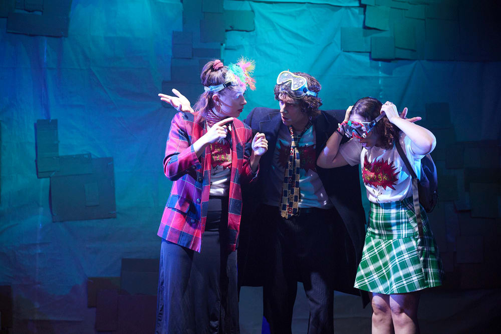 Image shows 3 people discussing something intently. They are stood against a green-lit backdrop. One is wearing a red checked coat, one is wearing a black suit jacket and the other is wearing a green skirt. All have goggles on their head.