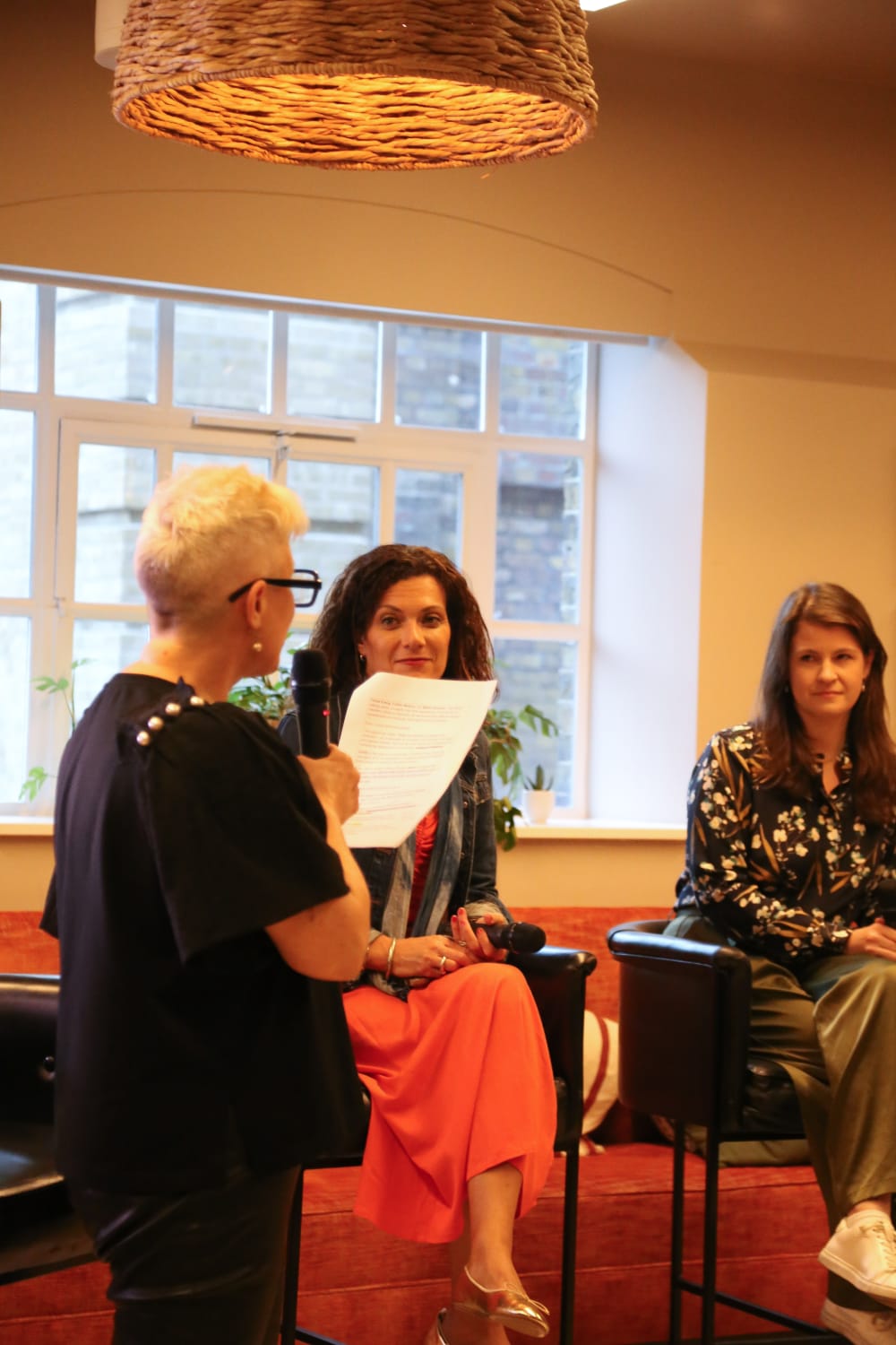 Two woman are on spotlight chairs while a host asks them questions from a sheet of paper