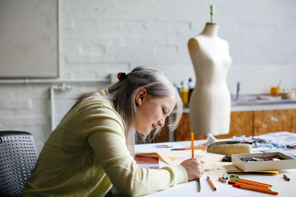 A female student with light purple hair and a green cardigan drawing some fashion illustrations with coloured pencils which are splayed across the table. There is a mannequin in the background. 