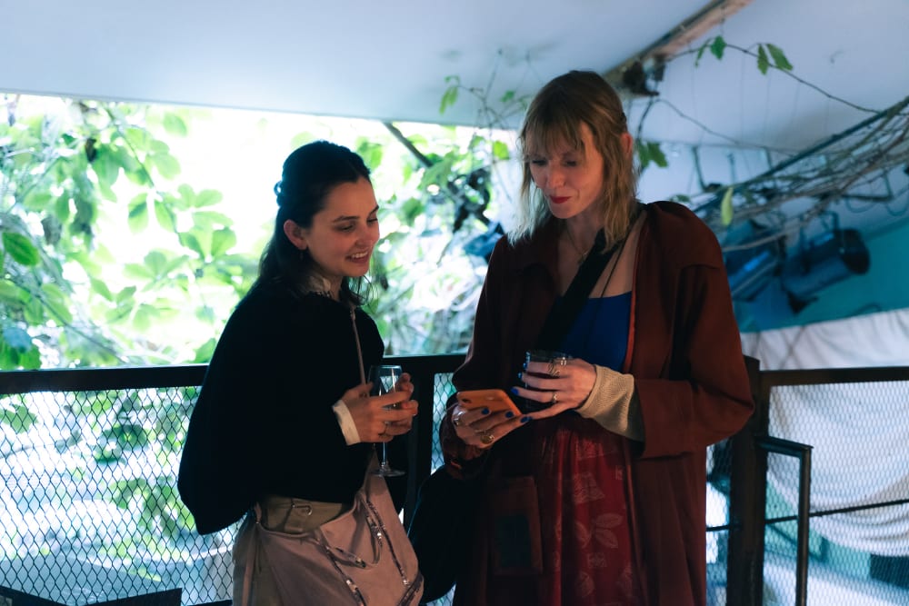 2 guests stood together looking at a phone. Behind them is a large window with plants growing over it.