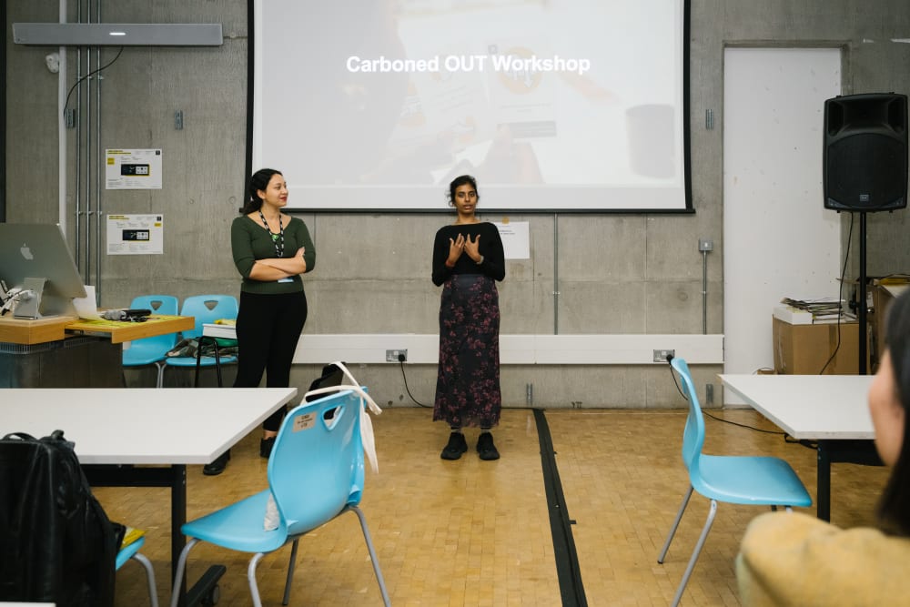 Ozge and Nanthini presenting in front of the room. Behind them on a screen it says 'Carboned Out workshop'