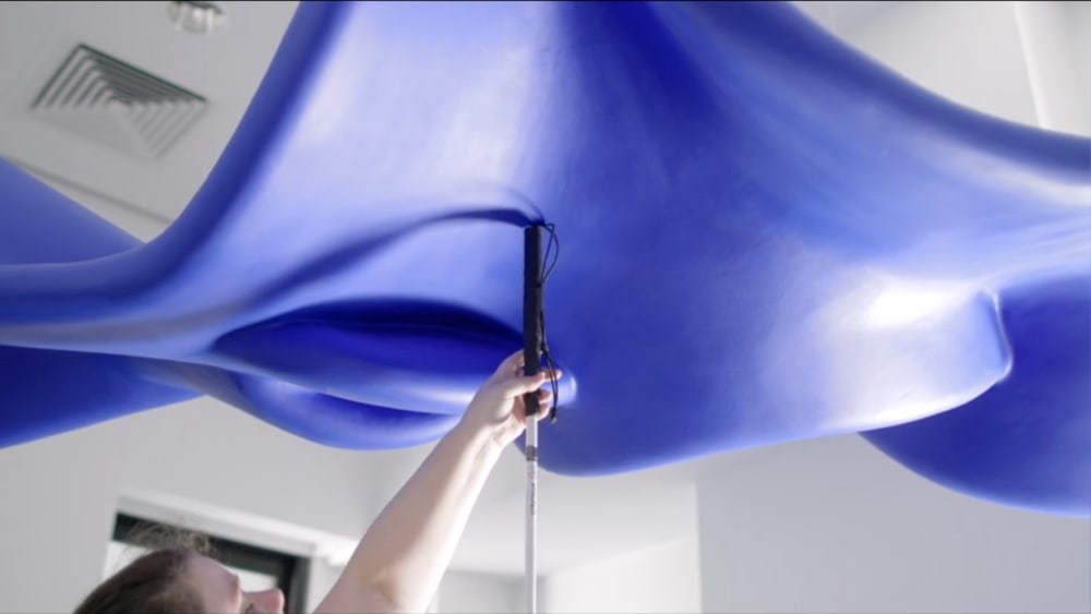 A person holds a cane, pointing it upwards at an undulating blue sculpture.