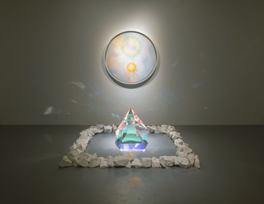 Image shows a large crystal sitting on the floor surrounded by white rocks, with a circular framed piece of artwork hanging on the wall behind. The crystal is reflecting light across the room, including onto the frame hanging on the wall.