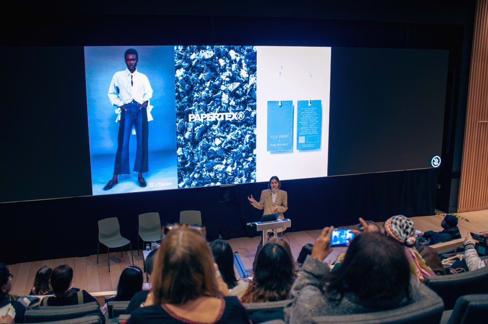 Wide shot of Kaela speaking, with guests seated in front of her. On the screen behind her are 3 images; one the left, a model in a white shirt and jeans with their hands in their pockets; in the middle a logo for Papertex; and on the right 2 clothing labels. 