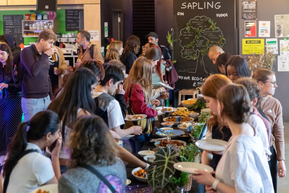 Guests queuing around the buffet of food. There are plants and greenery amongst the food to decorate the table.