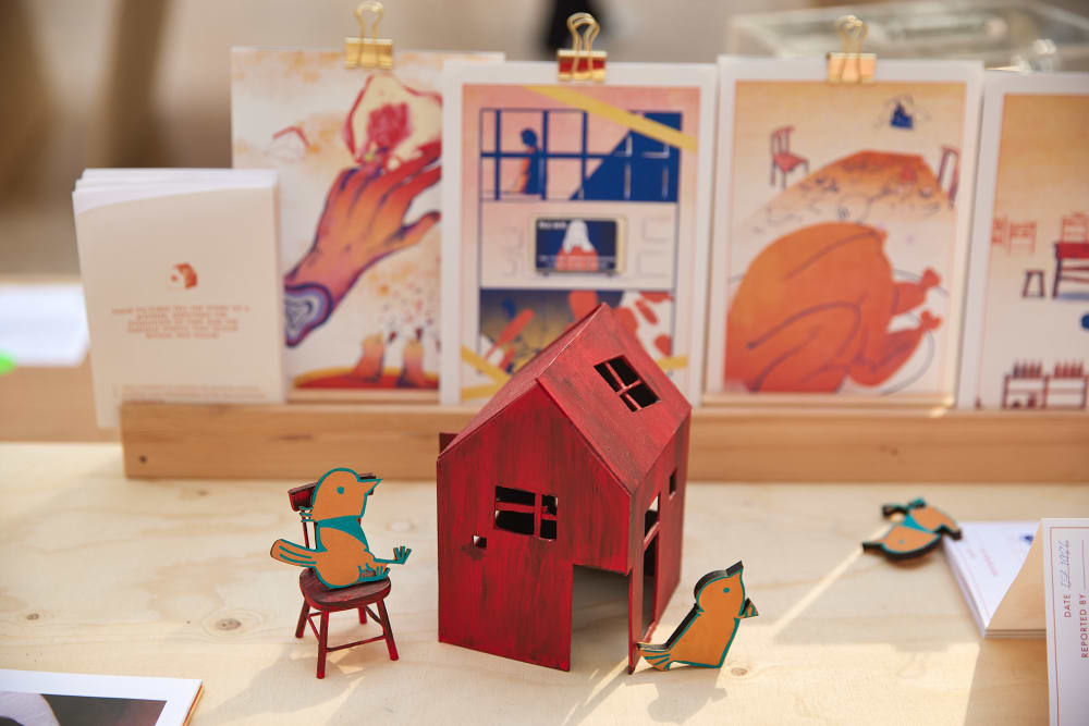 a small red-painted wooden house with wooden bird characters sitting on chairs and scattered around, with printed publications in the background.