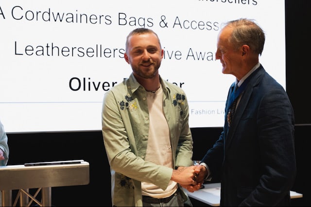 Two men shaking hands one with a grey shirt and blue jeans the other ina navy blue suit.