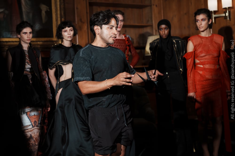 A man gestures while speaking, surrounded by five fashion models in elegant, avant-garde outfits. The setting is a dimly-lit, wood-paneled room, creating a serious and sophisticated ambiance.