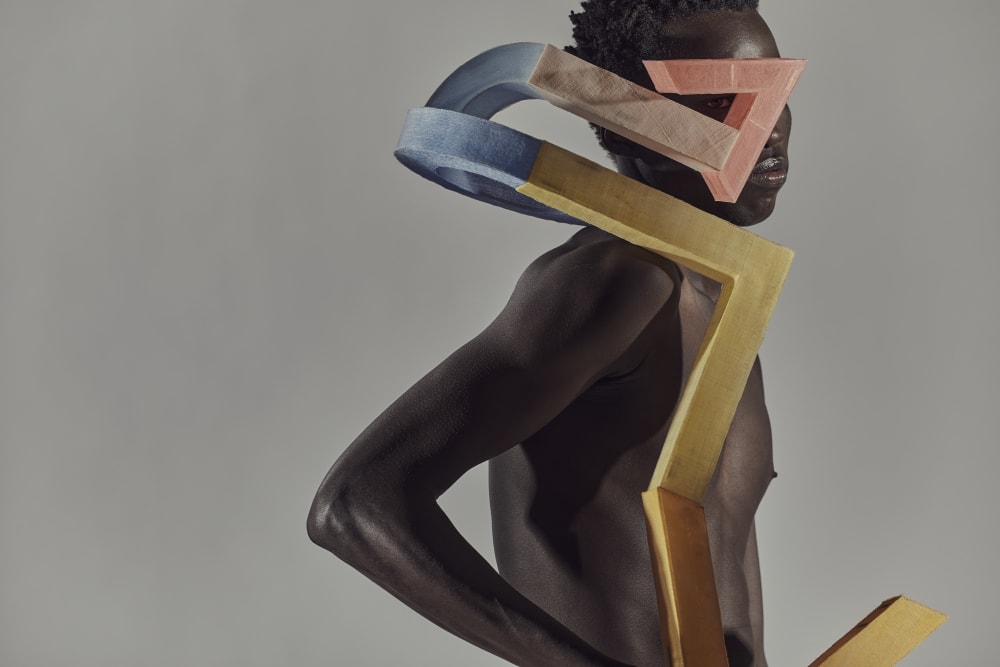 A shirtless person poses with angular, colorful geometric shapes on their head and body. The mood is artistic, abstract, and dynamic against a grey background.