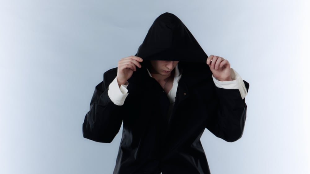 A person in a black hooded jacket partially conceals their face by holding the hood. The background is light blue, creating a mysterious, contemplative tone.