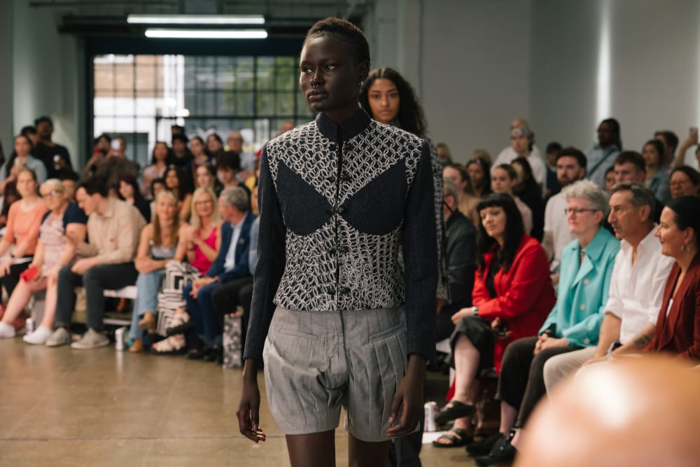 Model walking on a runway wearing a patterned top and gray shorts. The audience on both sides watches intently. The atmosphere is focused.
